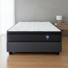A mattress on a dark gray upholstered bed base, photographed from the front against a plain white background. The mattress has a white quilted top and a black side panel with a small visible brand tag.