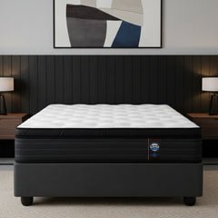 A mattress on a dark gray upholstered bed base, photographed from the front against a plain white background. The mattress has a white quilted top and a black side panel with a small visible brand tag.