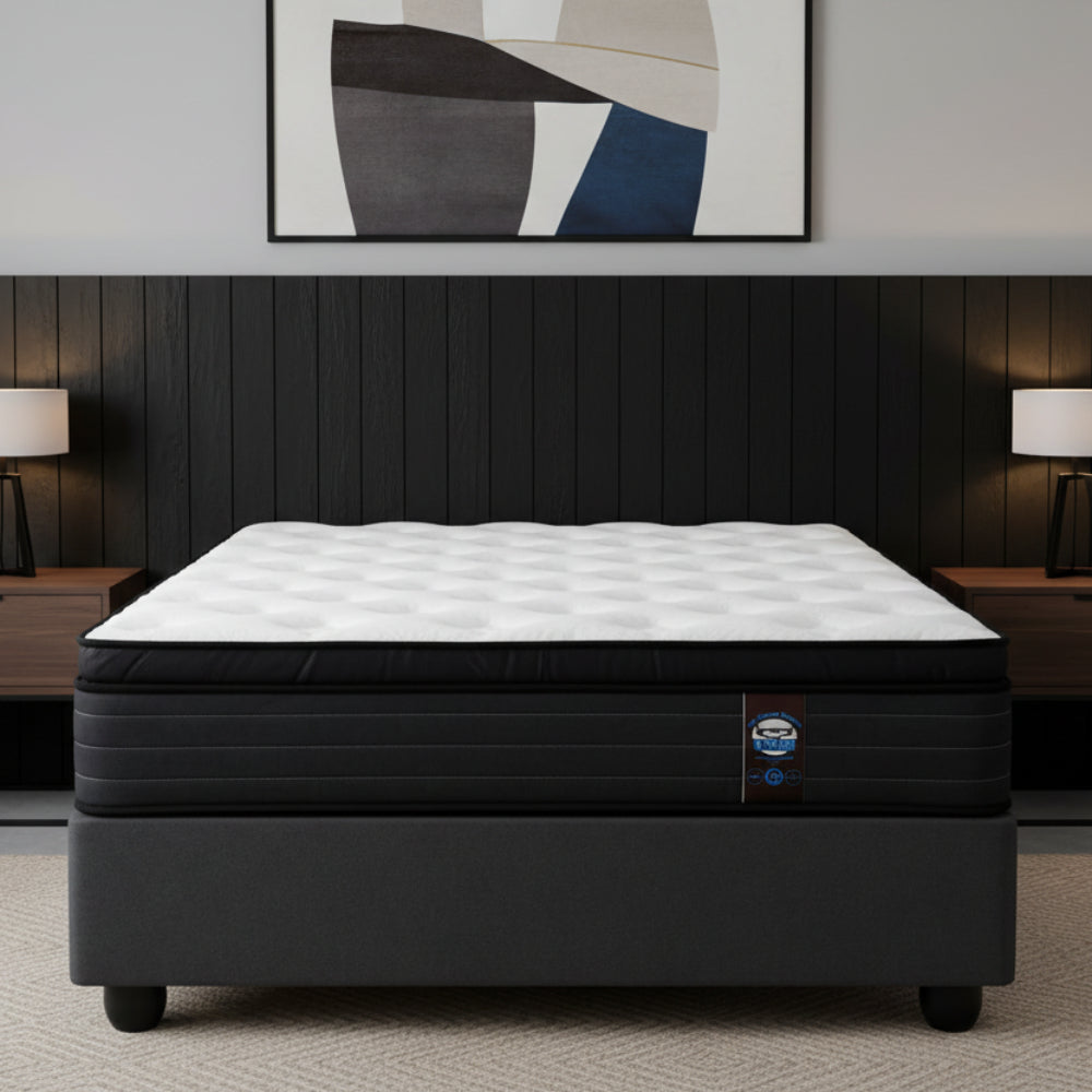 A mattress on a dark gray upholstered bed base, photographed from the front against a plain white background. The mattress has a white quilted top and a black side panel with a small visible brand tag.