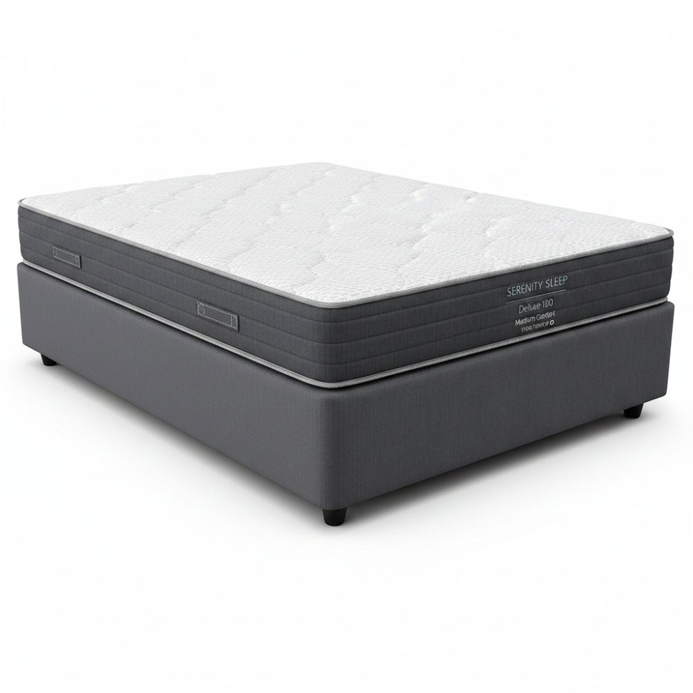 A Serenity Sleep Deluxe 100 medium comfort mattress on a dark gray upholstered box spring, shown from a front-facing perspective against a white background.