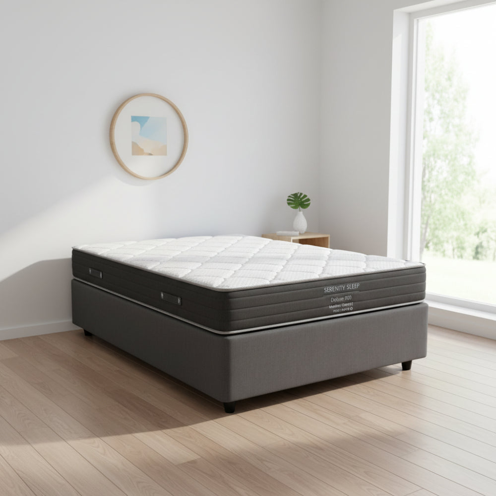 A Serenity Sleep Deluxe 100 medium comfort mattress on a dark gray upholstered box spring, shown from a front-facing perspective against a white background.
