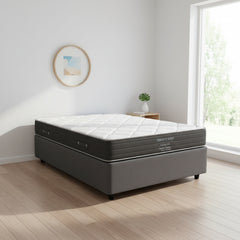 A Serenity Sleep Deluxe 100 medium comfort mattress on a dark gray upholstered box spring, shown from a front-facing perspective against a white background.