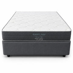 A Serenity Sleep Deluxe 100 medium comfort mattress on a dark gray upholstered box spring, shown from a front-facing perspective against a white background.