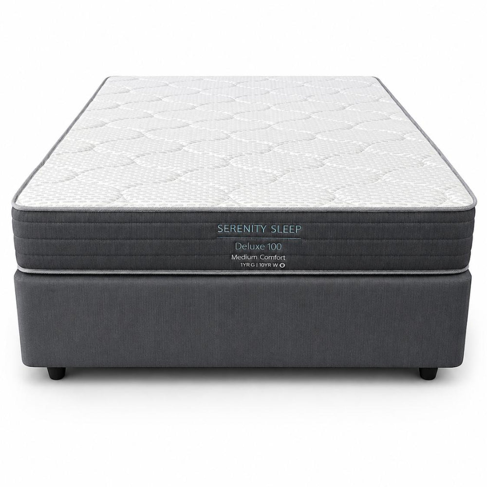 A Serenity Sleep Deluxe 100 medium comfort mattress on a dark gray upholstered box spring, shown from a front-facing perspective against a white background.
