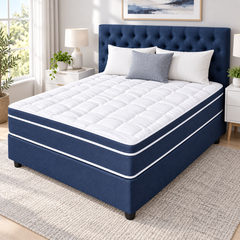 A bed with a navy blue tufted box spring and a white quilted mattress topper, styled in a bright bedroom with white and gray pillows, bedside tables, and a window.